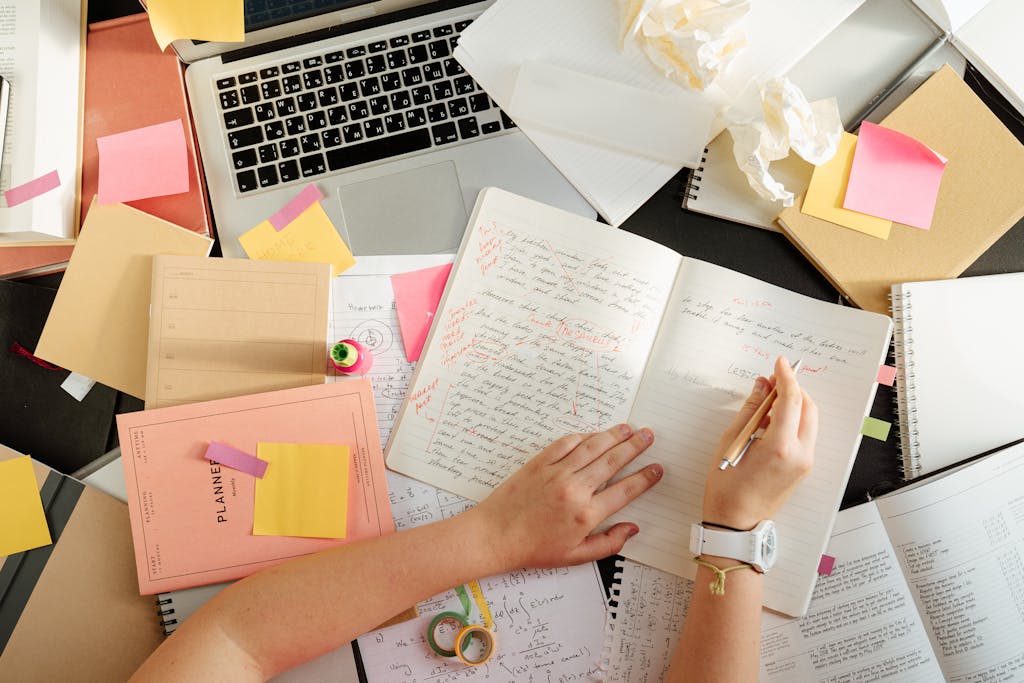 Overhead shot of a person organizing notes and study materials on a cluttered desk with a laptop.