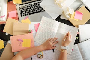 Overhead shot of a person organizing notes and study materials on a cluttered desk with a laptop.