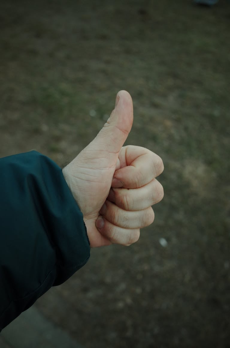 A close-up of a hand showing a thumbs up gesture in an outdoor park setting.