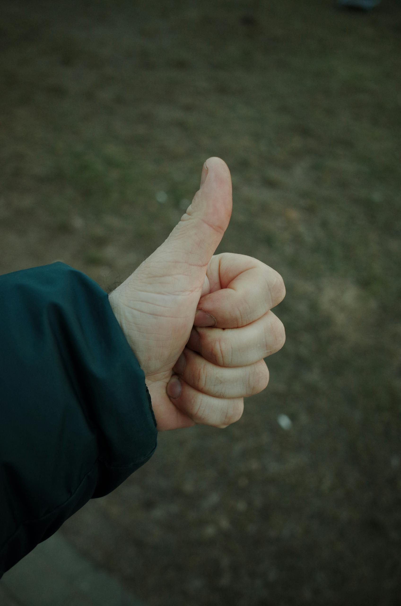 A close-up of a hand showing a thumbs up gesture in an outdoor park setting.