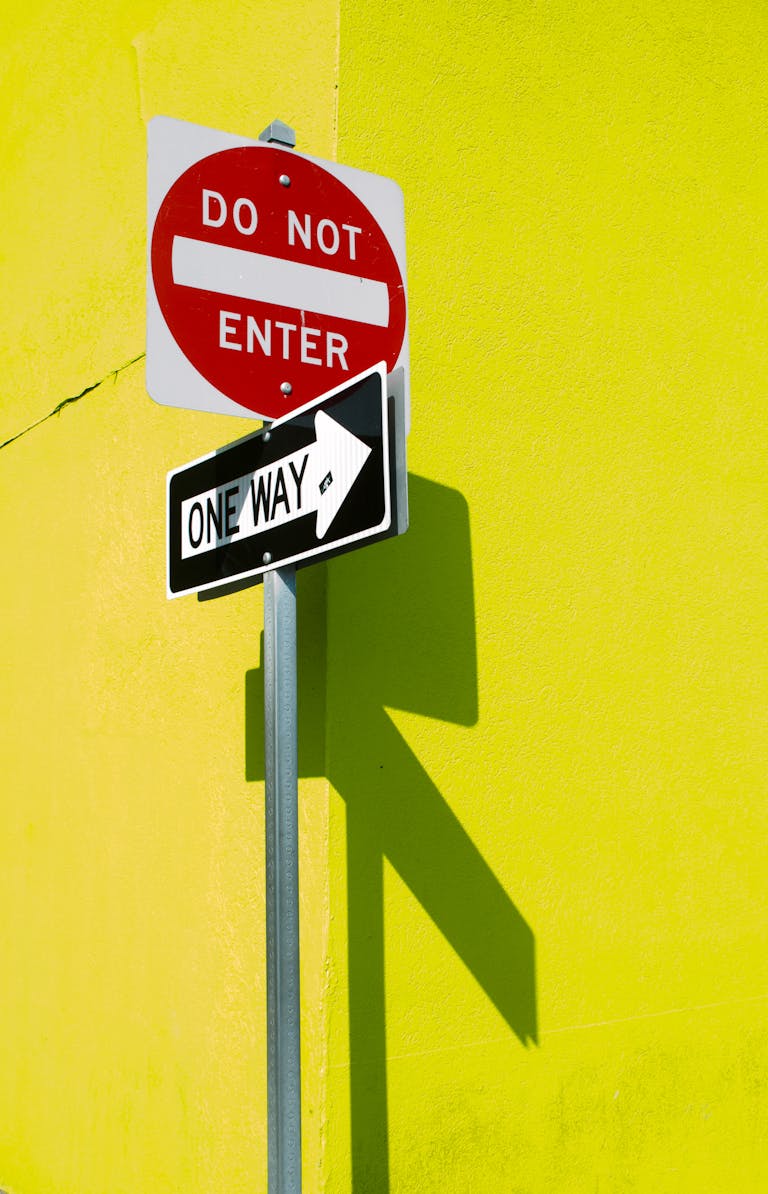 A vibrant yellow wall featuring a 'Do Not Enter' and 'One Way' sign casting shadows.