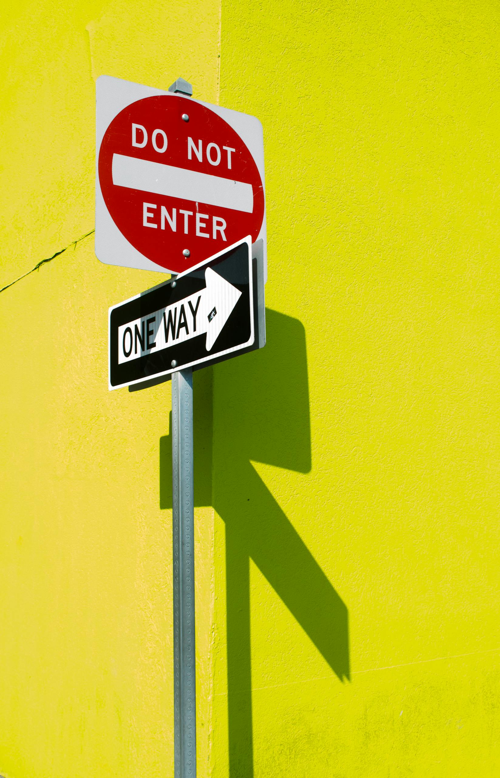 A vibrant yellow wall featuring a 'Do Not Enter' and 'One Way' sign casting shadows.