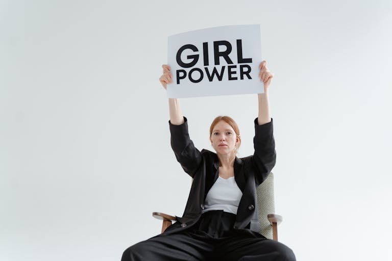 Confident woman in blazer sits with 'Girl Power' sign on white background.