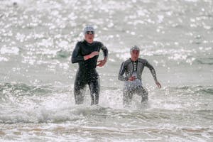 Focused young people in wet suits and swim caps running out of wavy glistening sea in sunlight
