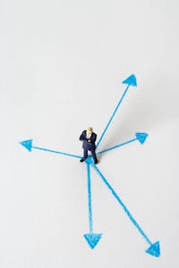Miniature businessman stands at crossroads of blue arrows on white background, symbolizing choices.