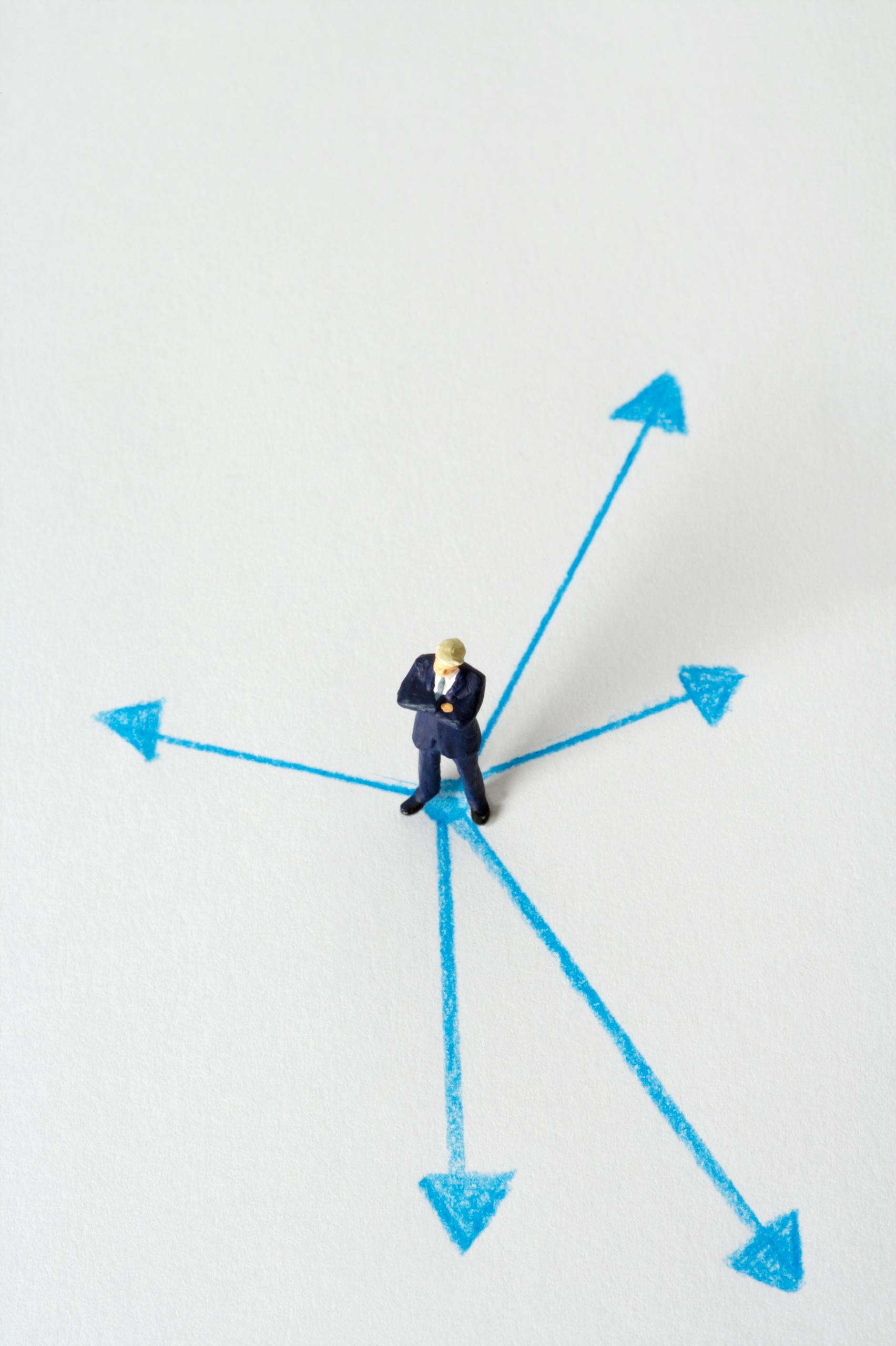 Miniature businessman stands at crossroads of blue arrows on white background, symbolizing choices.