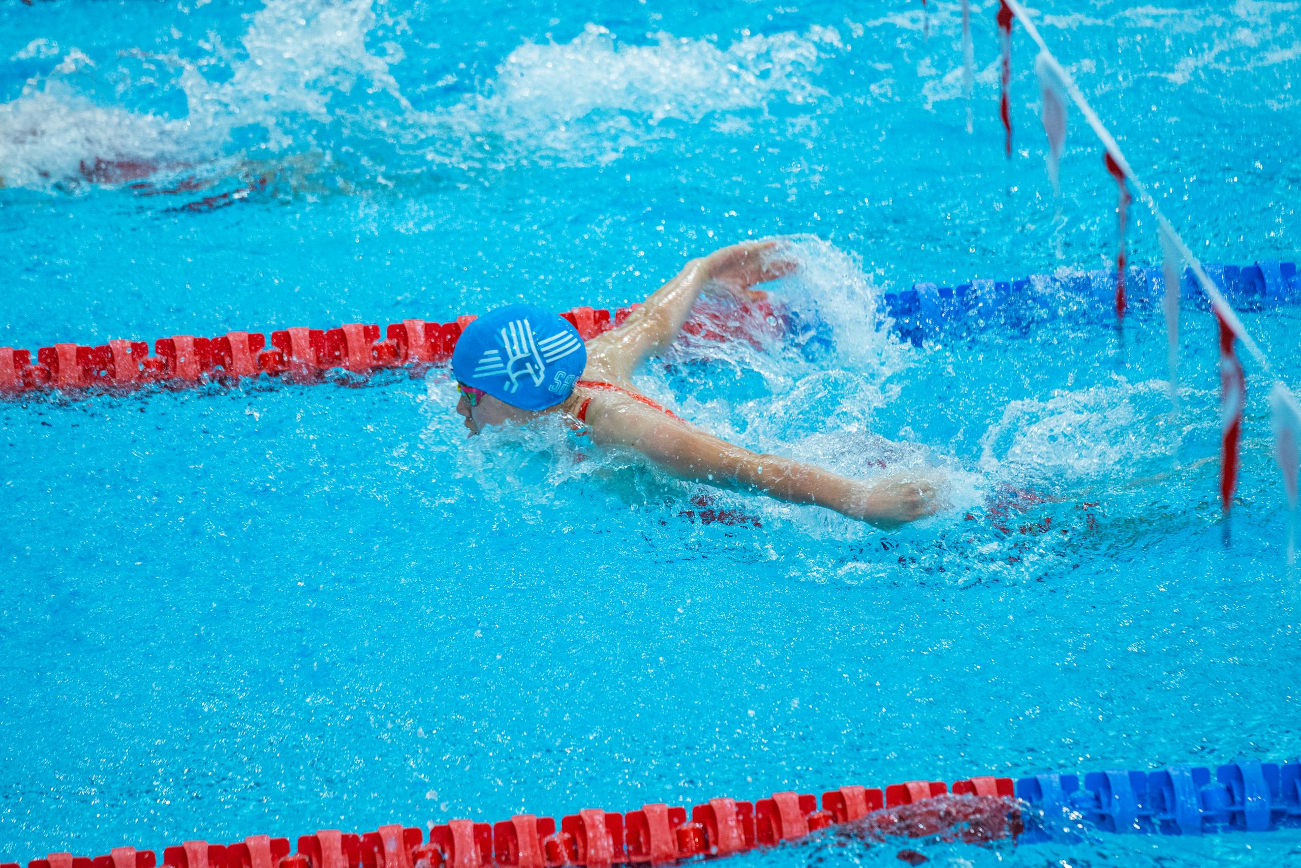 Swimmer in blue cap mid-butterfly stroke in indoor pool with red lane markers.